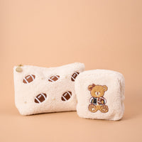 Add a playful touch to your collection with The Darling Effect's Square Touchdown Teddy Teddy Pouches—two soft sherpa pouches in creamy beige, including one embroidered with footballs and pearl accents, and another showcasing an adorable teddy bear holding a football.