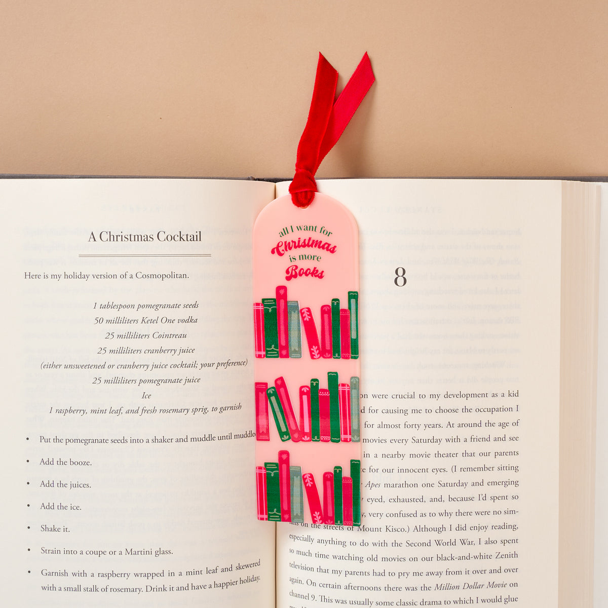 An All I Want for Christmas is More Books Acrylic Bookmark, featuring a pale pink design with a velvet ribbon topper and colorful book illustrations, resting in an open book next to a cocktail recipe.