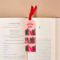 An All I Want for Christmas is More Books Acrylic Bookmark, featuring a pale pink design with a velvet ribbon topper and colorful book illustrations, resting in an open book next to a cocktail recipe.