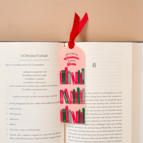 An All I Want for Christmas is More Books Acrylic Bookmark, featuring a pale pink design with a velvet ribbon topper and colorful book illustrations, resting in an open book next to a cocktail recipe.