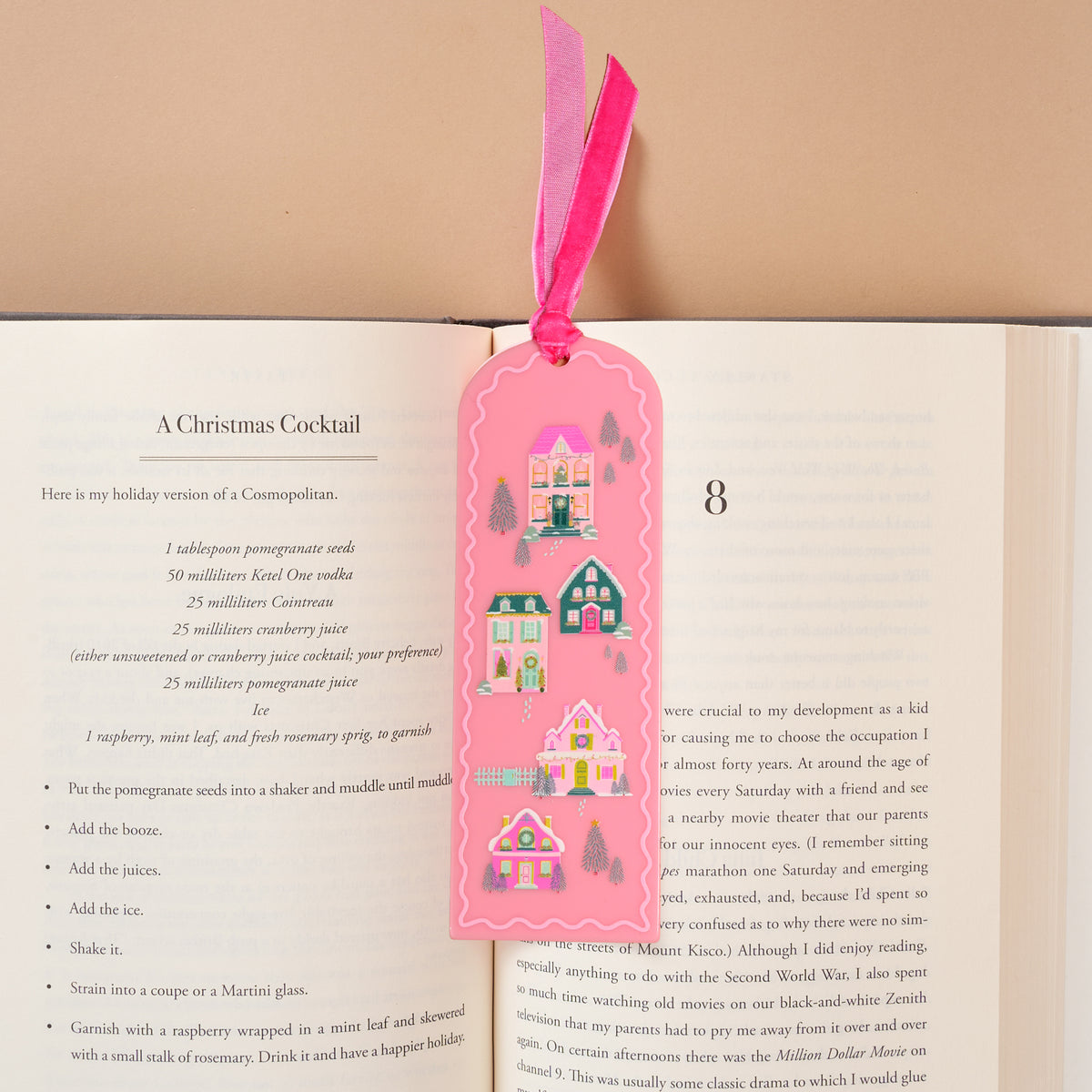 Pink Pine Village Acrylic Bookmark by The Darling Effect, featuring a scalloped pink shape decorated with illustrations of colorful houses, pine trees, and a deer, tucked into a book with a pink velvet ribbon topper.