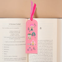Pink Pine Village Acrylic Bookmark by The Darling Effect, featuring a scalloped pink shape decorated with illustrations of colorful houses, pine trees, and a deer, tucked into a book with a pink velvet ribbon topper.