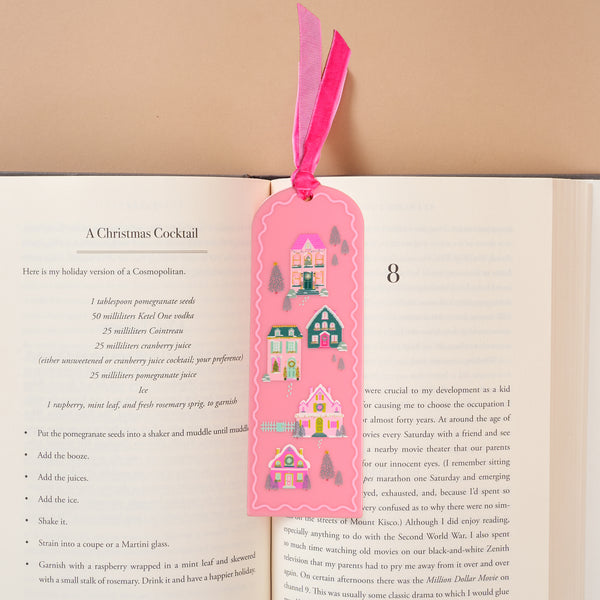Pink Pine Village Acrylic Bookmark by The Darling Effect, featuring a scalloped pink shape decorated with illustrations of colorful houses, pine trees, and a deer, tucked into a book with a pink velvet ribbon topper.