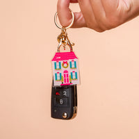 A hand holds a gold keyring with a BowBelle Manor Acrylic Keychain shaped like a house with a pink roof and green wreath, attached to a black car key fob.