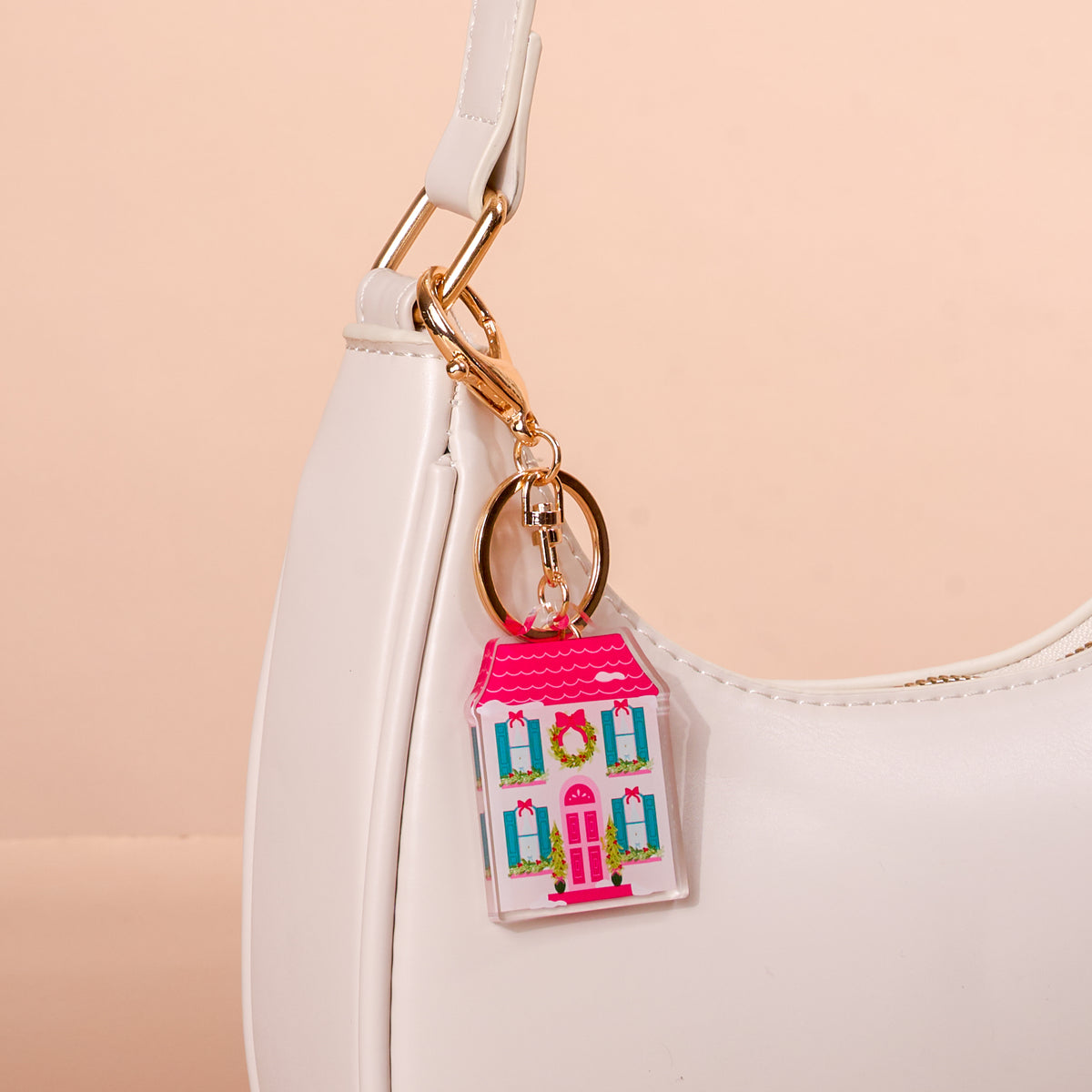 A BowBelle Manor Acrylic Keychain shaped like a pink house with green wreaths on the windows, attached to the gold hardware of a white handbag.