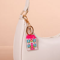 A BowBelle Manor Acrylic Keychain shaped like a pink house with green wreaths on the windows, attached to the gold hardware of a white handbag.
