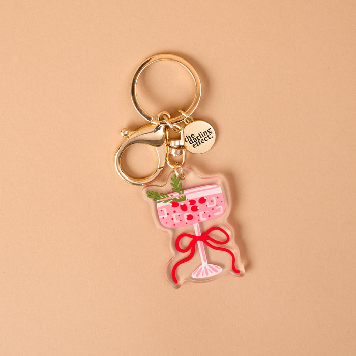 Cranberry Cosmo Acrylic Keychain shaped like a pink cocktail in a stemmed glass, garnished with a sprig of rosemary and cranberries, and tied with a red ribbon.
