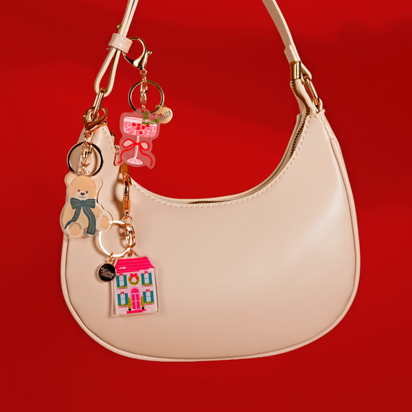 A beige crescent handbag decorated with a Cranberry Cosmo Acrylic Keychain alongside two other acrylic charms shaped like a teddy bear and a house.