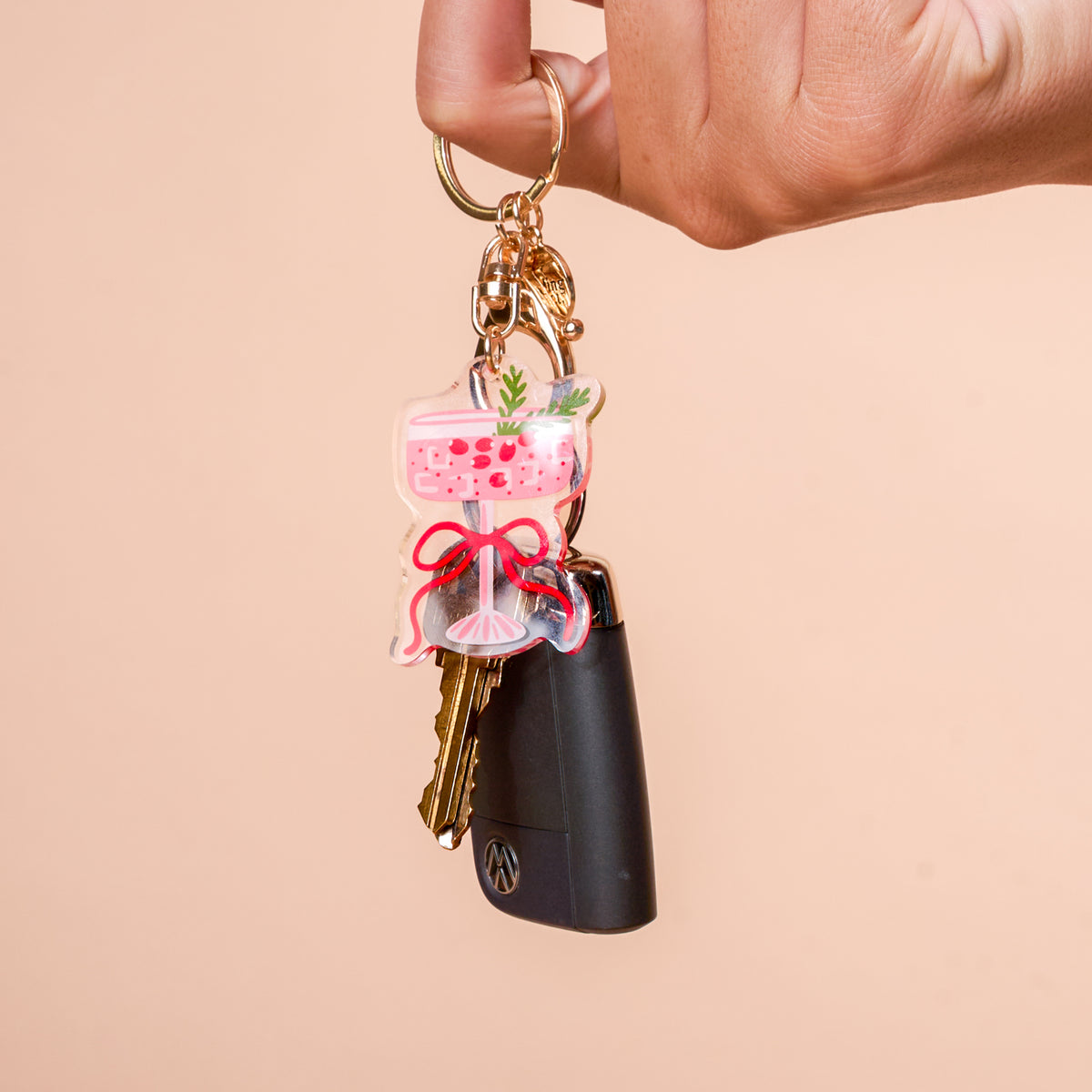 A hand holds a car key with a Cranberry Cosmo Acrylic Keychain by The Darling Effect, featuring a pink cocktail in a coupe glass decorated with a red ribbon and a sprig of greenery.