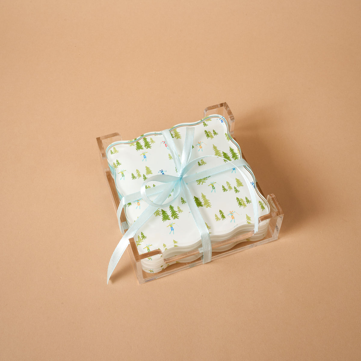 The Aprés Ski Table Talk Acrylic Coaster Set features square, scalloped-edge coasters with a pine tree and skier pattern, tied with a blue ribbon and displayed in a clear acrylic stand as stylish home décor.