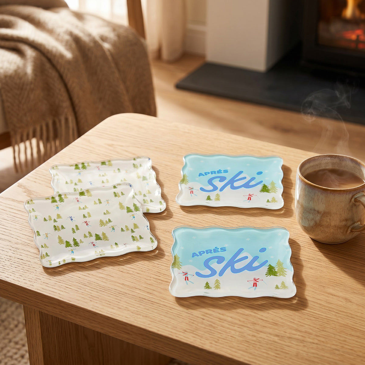 Four Aprés Ski Table Talk Acrylic Coaster Set coasters on a wooden table, including two with a forest print and two featuring the text Après ski with illustrated skiers.