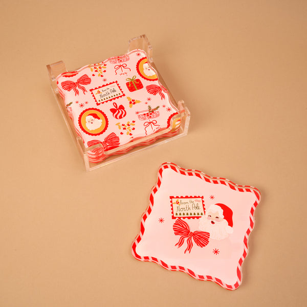 A Mistletoe Magic Table Talk Acrylic Coaster Set of four square coasters with wavy edges in a clear holder, featuring Christmas illustrations of Santa Claus, gift boxes, and the text From the North Pole.
