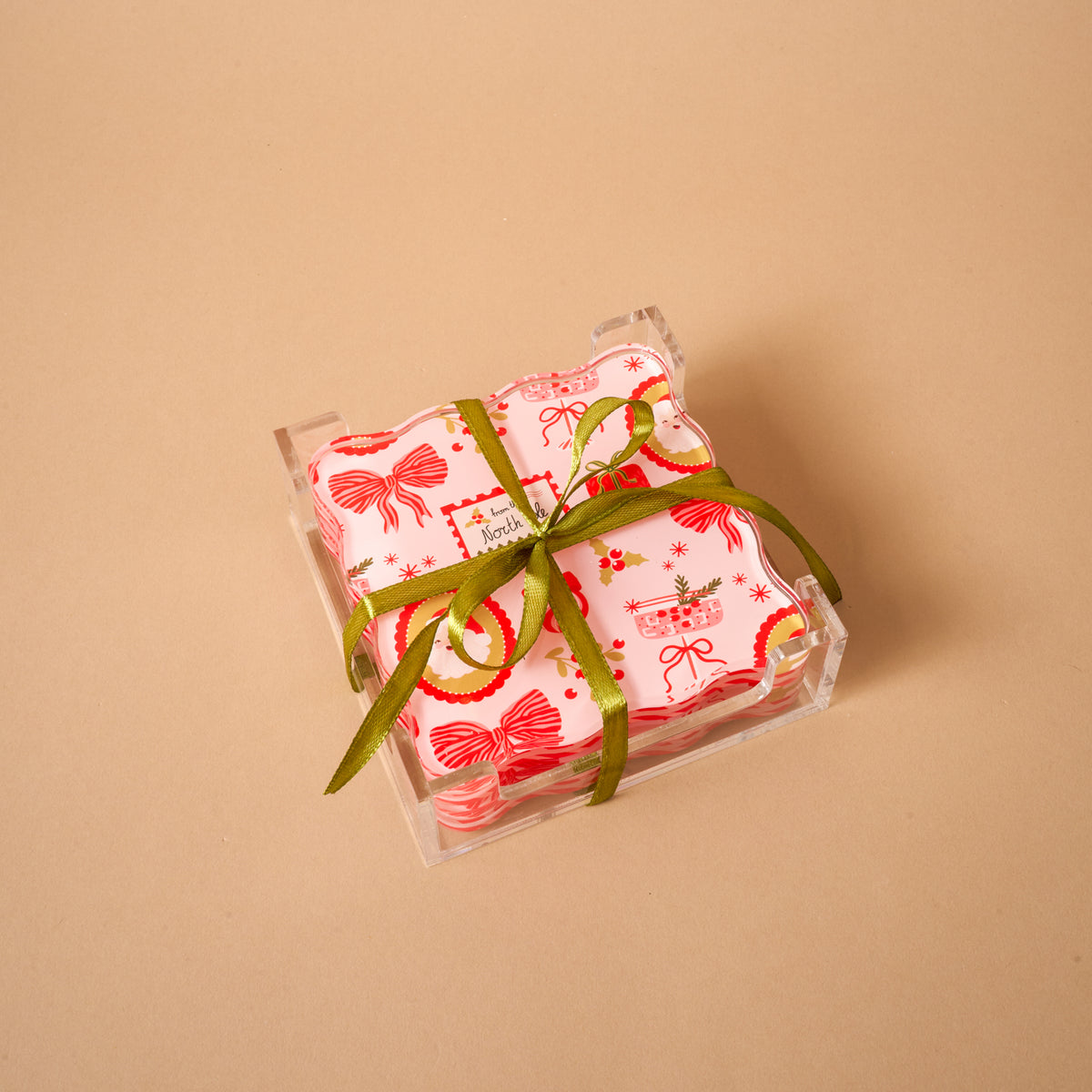 Mistletoe Magic Table Talk Acrylic Coaster Set featuring square pink coasters with festive red and gold Christmas patterns, tied with a green ribbon in a clear holder, for stylish home décor.