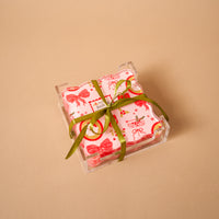 Mistletoe Magic Table Talk Acrylic Coaster Set featuring square pink coasters with festive red and gold Christmas patterns, tied with a green ribbon in a clear holder, for stylish home décor.