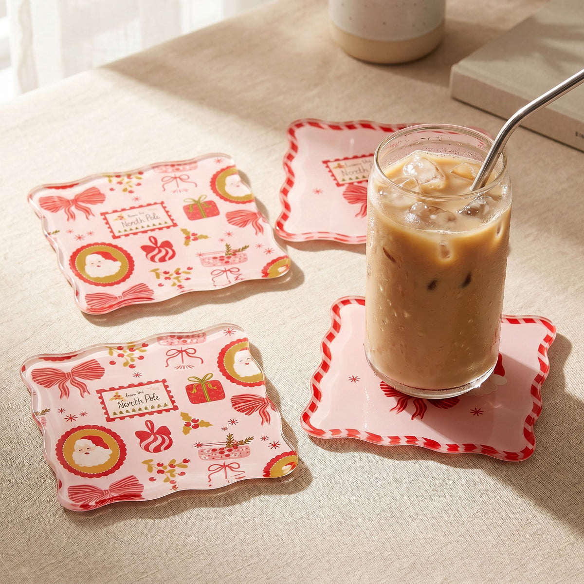 Three square Mistletoe Magic Table Talk Acrylic Coaster Set coasters decorated with Christmas icons like Santa, gifts, and bows, with an iced coffee sitting on one piece of artistic home décor.