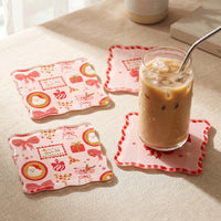 Three square Mistletoe Magic Table Talk Acrylic Coaster Set coasters decorated with Christmas icons like Santa, gifts, and bows, with an iced coffee sitting on one piece of artistic home décor.