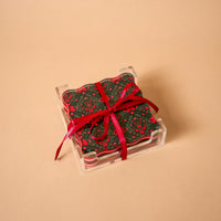 A Merry Moment Table Talk Acrylic Coaster Set featuring square, dark green acrylic coasters with red floral patterns, tied with a red ribbon and displayed in a clear holder for elegant home décor.