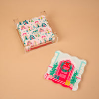 A stack of Sugarplum Lane Table Talk Acrylic Coaster Set square coasters in a clear holder, with one matching coaster featuring a festive house pattern, pink door, and Christmas wreath as artistic home décor.