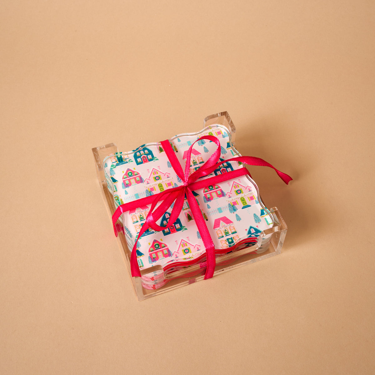 A Sugarplum Lane Table Talk Acrylic Coaster Set featuring festive village prints, stacked in a clear holder and tied with a bright pink ribbon as functional home décor.