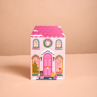The Adoring Ornaments Ornament Advent Calendar is a pink, house-shaped ornament calendar decorated with holiday wreaths, Christmas trees, and wrapped gifts.