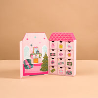 The Adoring Ornaments Ornament Advent Calendar is a house-shaped advent calendar with twelve numbered drawers and a decorative interior illustration of a cozy living room with a Christmas tree and fireplace, designed for miniature acrylic ornaments.