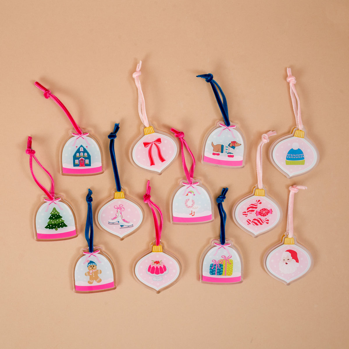Twelve bell-shaped miniature acrylic ornaments from the Adoring Ornaments Ornament Advent Calendar, featuring various holiday illustrations and colored ribbon ties, arranged on a tan background.