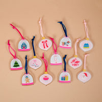 Twelve bell-shaped miniature acrylic ornaments from the Adoring Ornaments Ornament Advent Calendar, featuring various holiday illustrations and colored ribbon ties, arranged on a tan background.