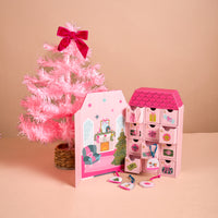 A pink, house-shaped Adoring Ornaments Ornament Advent Calendar with twelve numbered drawers stands next to a small pink artificial Christmas tree, with miniature acrylic ornaments spread on the surface in front.