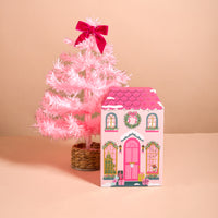 A pink artificial Christmas tree with a bright pink bow stands next to an Adoring Ornaments Ornament Advent Calendar, a house-shaped advent calendar illustrated with windows, a front door, and holiday décor.