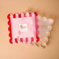 A Peppermint Stripes Square Scalloped Acrylic Picture Frame featuring red and pink vertical stripes stacked with a translucent square scalloped acrylic frame against a tan background.