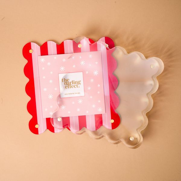 A Peppermint Stripes Square Scalloped Acrylic Picture Frame featuring red and pink vertical stripes stacked with a translucent square scalloped acrylic frame against a tan background.