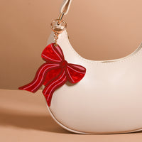 A glossy red Red Bow Mini Mirror Bag Charm by The Darling Effect attached to the strap of a cream-colored crescent bag.