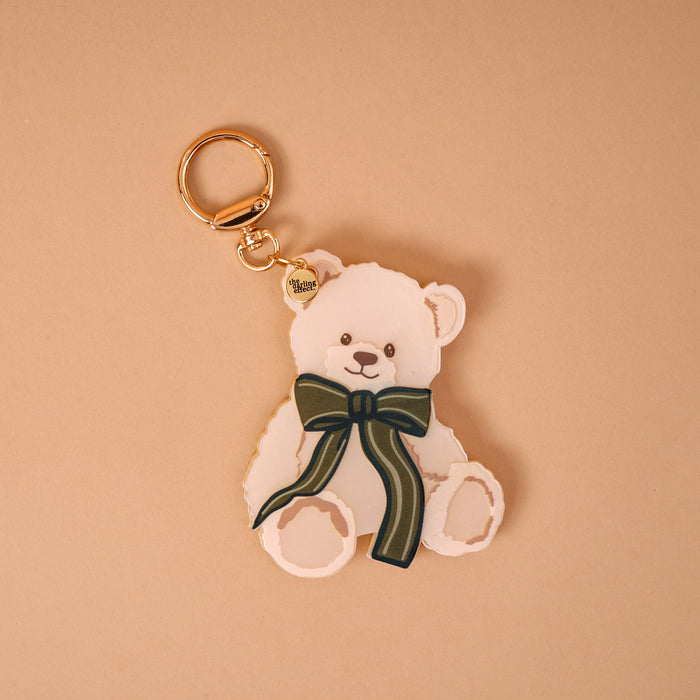 Teddy Bear Theo Mini Mirror Bag Charm, a light-colored bear-shaped acrylic mirror featuring a dark green striped bow and a gold-tone keyring.