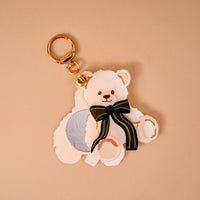 Teddy Bear Theo Mini Mirror Bag Charm in the shape of a teddy bear-shaped acrylic mirror, featuring a small circular mirror and a green striped bow, attached to a gold key ring.