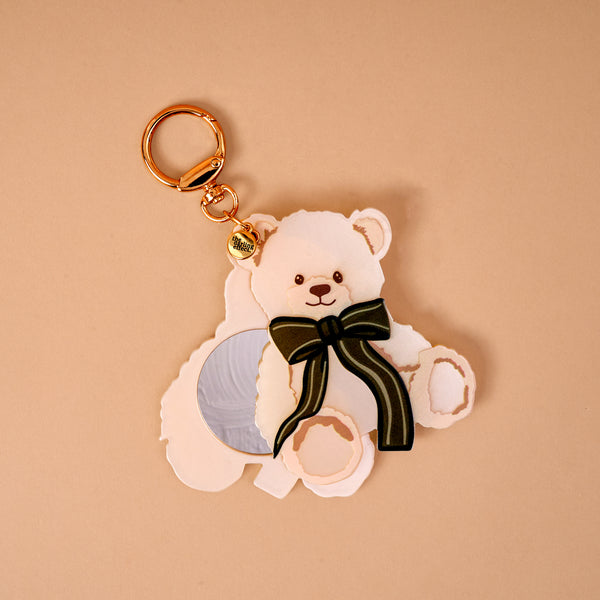 Teddy Bear Theo Mini Mirror Bag Charm in the shape of a teddy bear-shaped acrylic mirror, featuring a small circular mirror and a green striped bow, attached to a gold key ring.