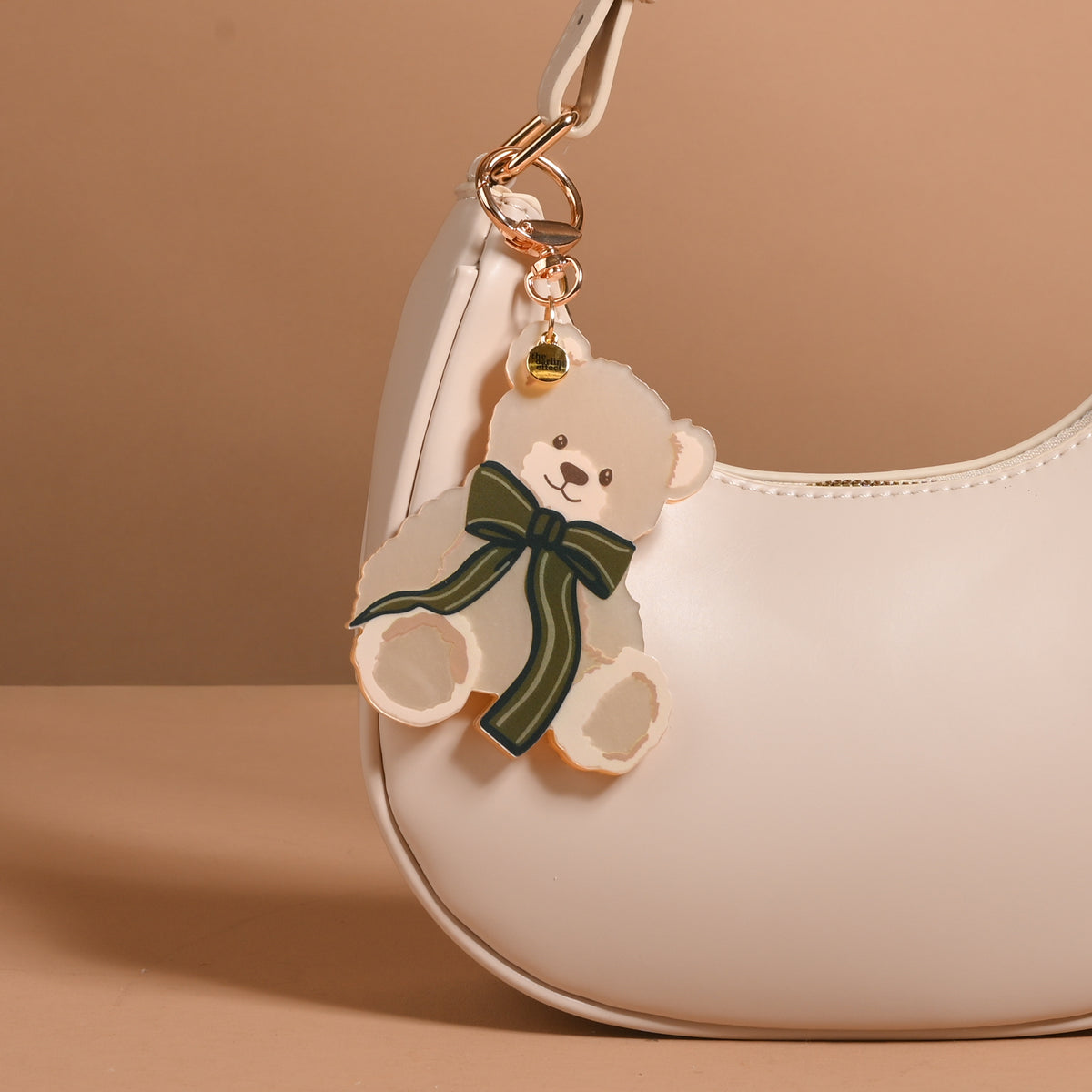 A Teddy Bear Theo Mini Mirror Bag Charm, featuring a teddy bear-shaped acrylic mirror with a dark green bow, attached to the gold hardware of a cream-colored handbag.