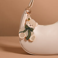A Teddy Bear Theo Mini Mirror Bag Charm, featuring a teddy bear-shaped acrylic mirror with a dark green bow, attached to the gold hardware of a cream-colored handbag.