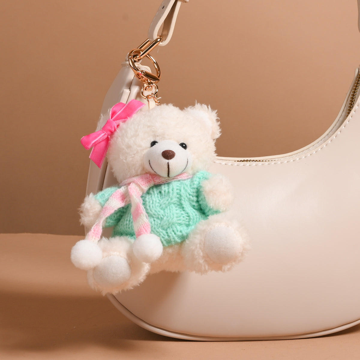 An Arctic Angel Teddy Bear Bag Charm wearing a green sweater, a pink and white striped scarf, and a pink hair bow, clipped to a cream-colored handbag.