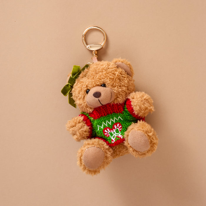 The Peppermint Twist Teddy Bear Bag Charm is a teddy bear wearing a green and red holiday sweater with a candy cane heart design and a small green hair bow, attached to a gold lobster clasp.