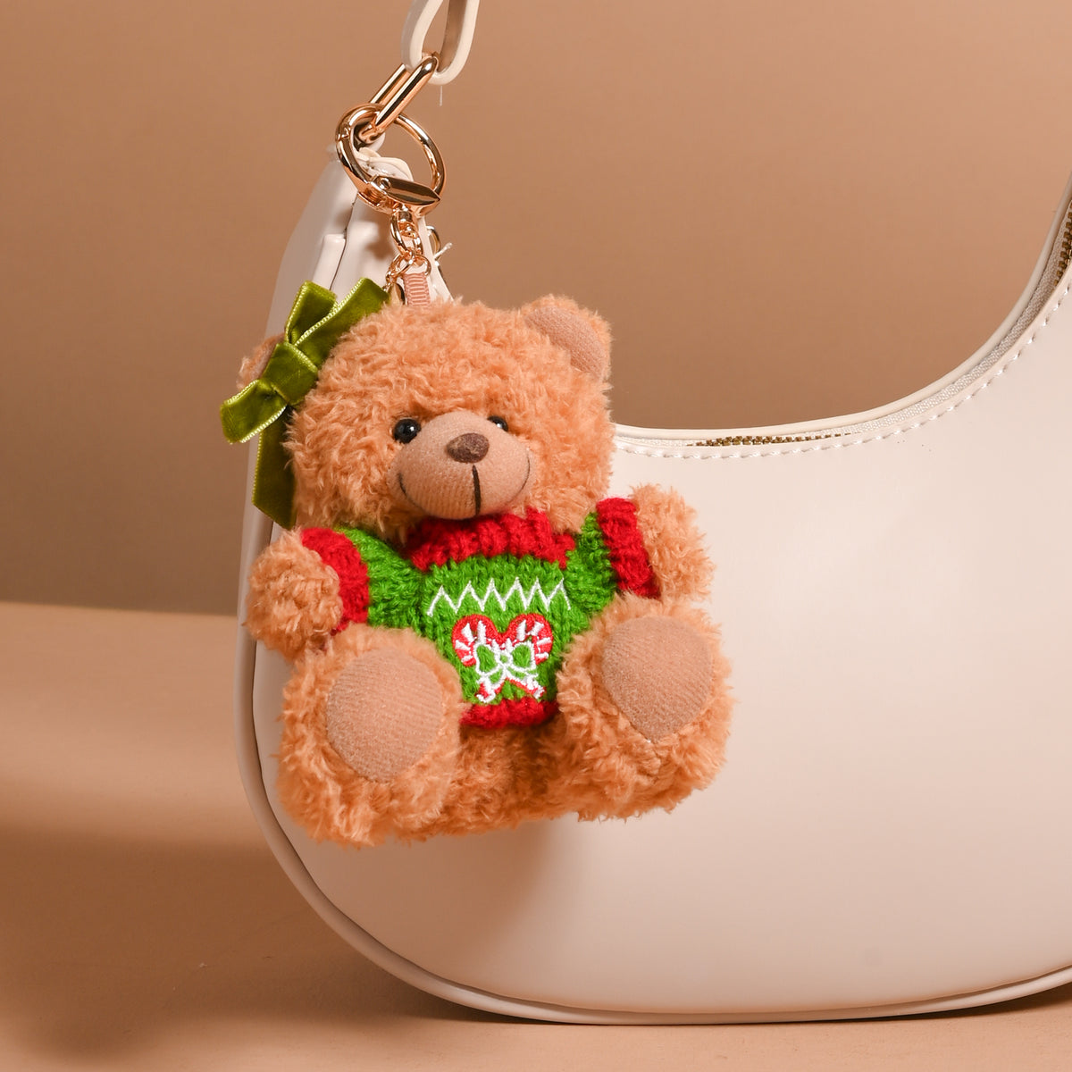 The Darling Effect Peppermint Twist Teddy Bear Bag Charm, a plush bear wearing a holiday peppermint sweater and a green ear bow, clipped to a white handbag as a festive accessory.