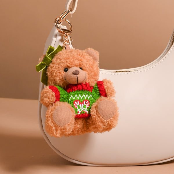 The Darling Effect Peppermint Twist Teddy Bear Bag Charm, a plush bear wearing a holiday peppermint sweater and a green ear bow, clipped to a white handbag as a festive accessory.