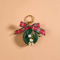 A small, circular, green Pine & Plaid Travel Jewelry Case bag charm by The Darling Effect, decorated with a plaid bow and gold teddy bear, Christmas tree, and pearl bead charms.