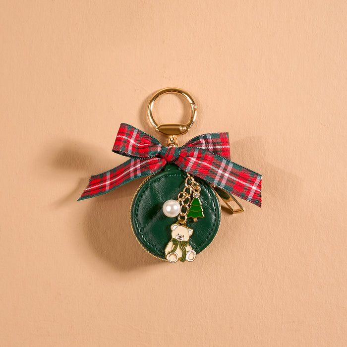 A small, circular, green Pine & Plaid Travel Jewelry Case bag charm by The Darling Effect, decorated with a plaid bow and gold teddy bear, Christmas tree, and pearl bead charms.