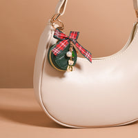 A green, circular Pine & Plaid Travel Jewelry Case bag charm hangs from a cream-colored handbag, decorated with a red plaid bow, a faux pearl, and a small gold bear pendant.