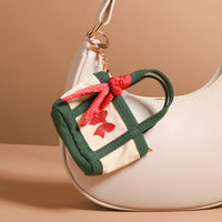 A Merry Little Bow Tiny Tote Bag Charm in cream and green with a red bow design, clipped to the strap of a larger cream shoulder bag.