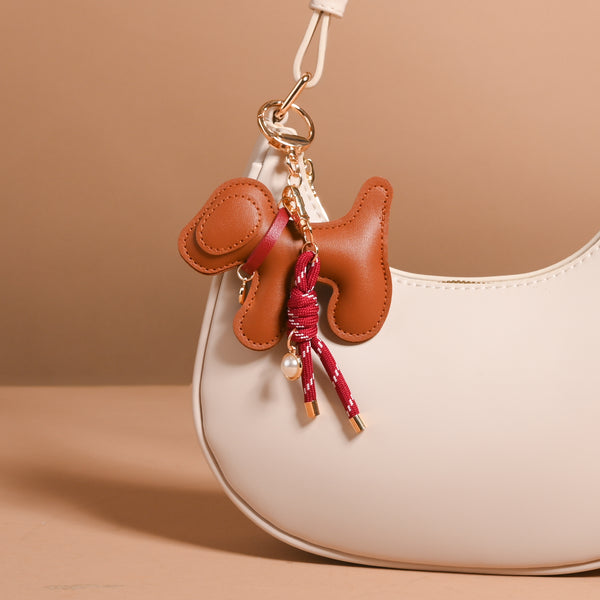 Posh Pup Vegan Leather Bag Charm shaped like a brown dog, featuring a red rope tassel and small pearl accent, hanging from the strap of a cream-colored handbag.
