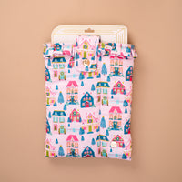 The Sugarplum Lane Book Cover is a padded, quilted book cover featuring a colorful pattern of houses, Christmas trees, and wreaths, secured with a ruffled flap and snap button for book protection.