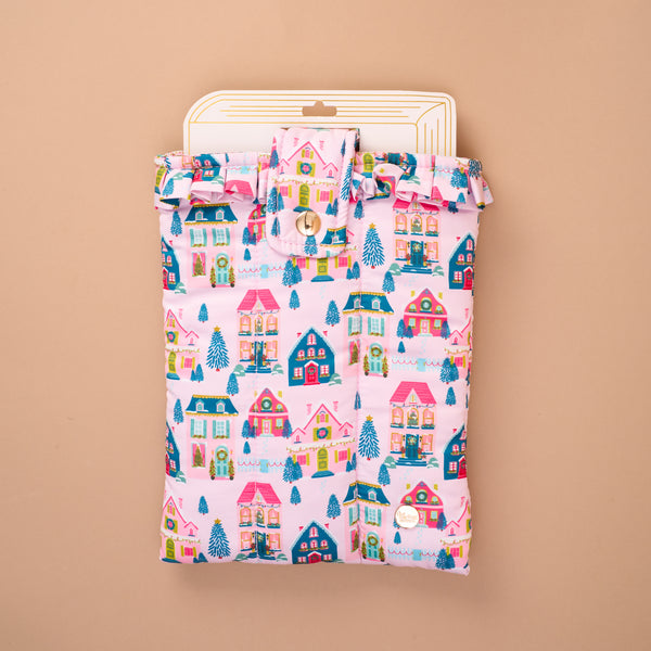 The Sugarplum Lane Book Cover is a padded, quilted book cover featuring a colorful pattern of houses, Christmas trees, and wreaths, secured with a ruffled flap and snap button for book protection.
