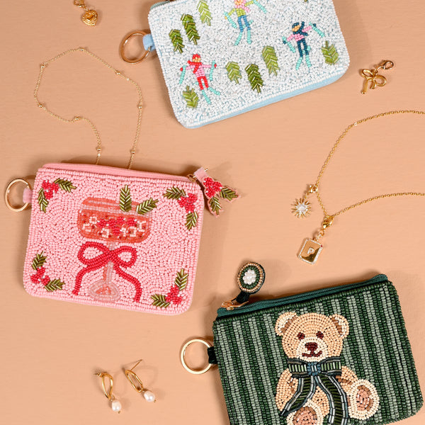 Three handcrafted beaded pouches, including the Cranberry Cosmo Handcrafted Beaded Pouch, alongside those featuring winter skiers and a teddy bear, arranged on a tan surface with gold jewelry.