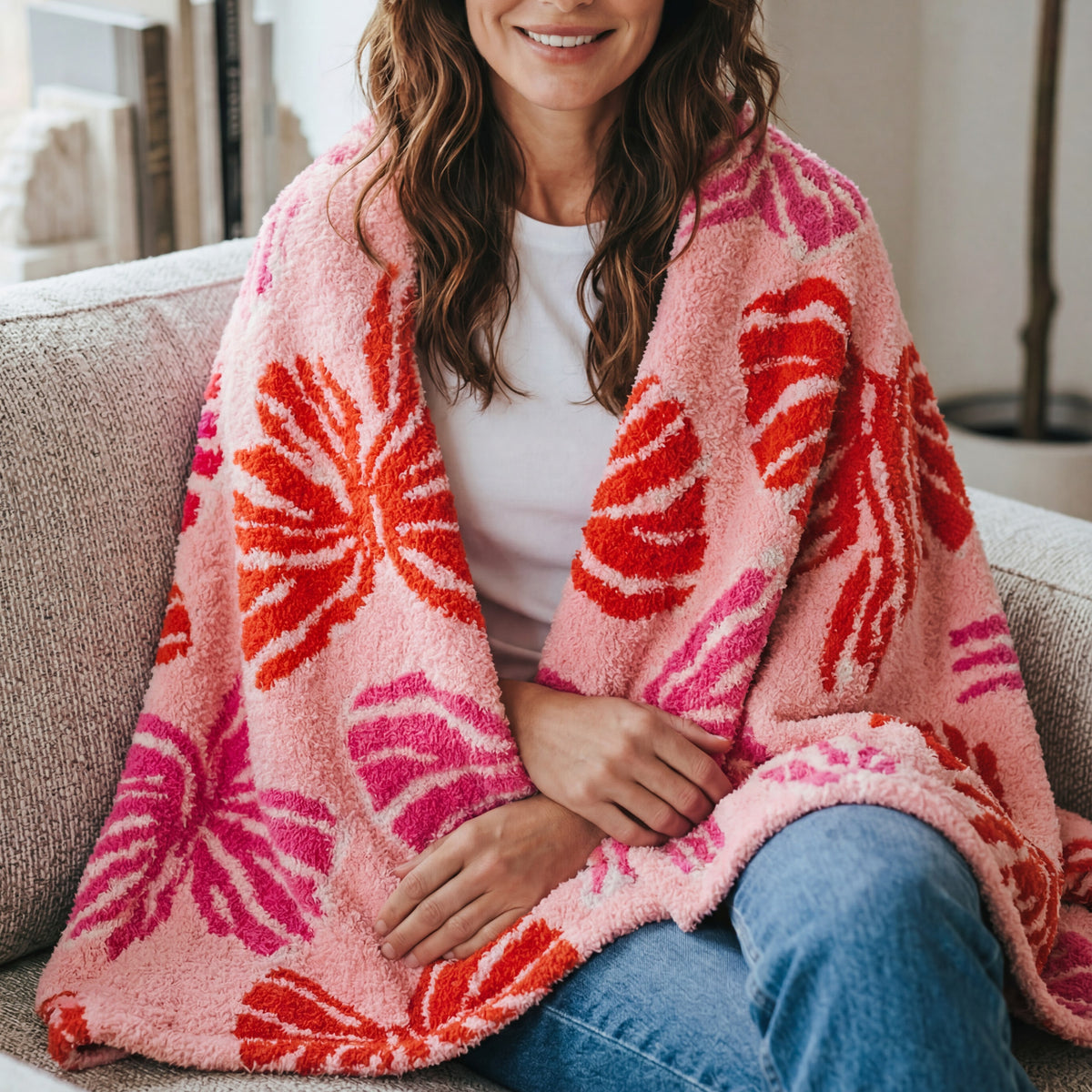 A person sits on a sofa draped in an ultra-soft Wrapped in Bows Cozy Dreams Luxe Blanket by The Darling Effect, featuring a large red and pink tropical leaf pattern.